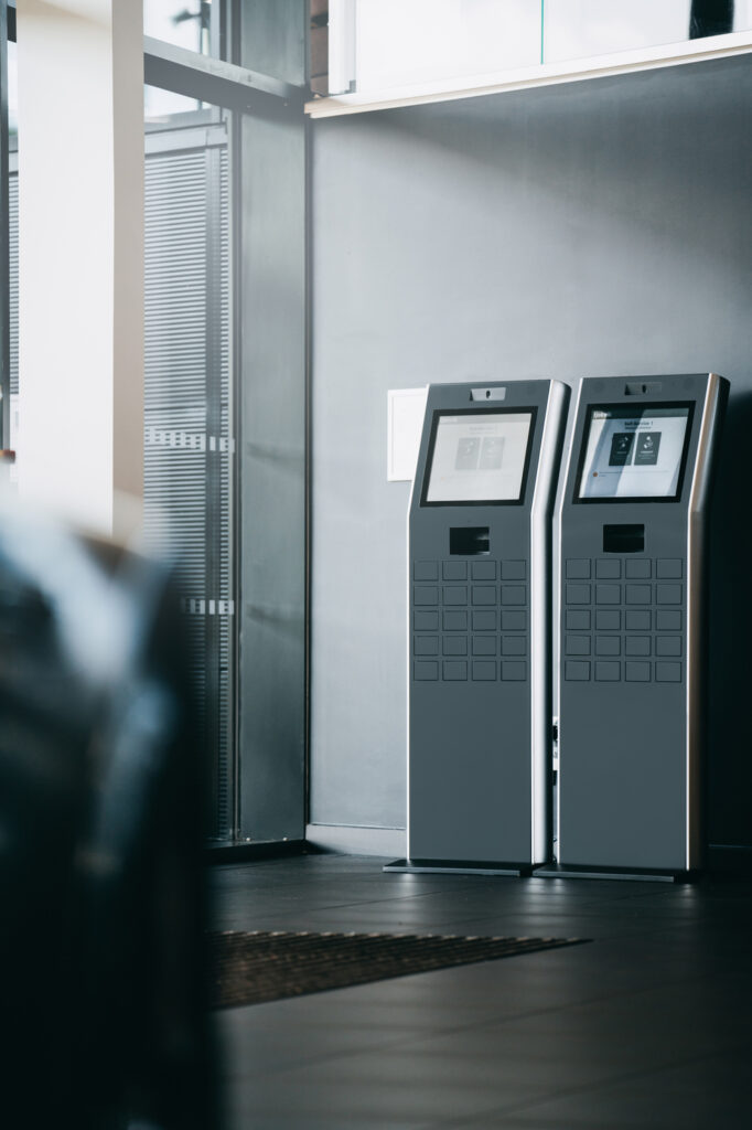 Two self-service kiosks with touchscreens in a modern lobby.