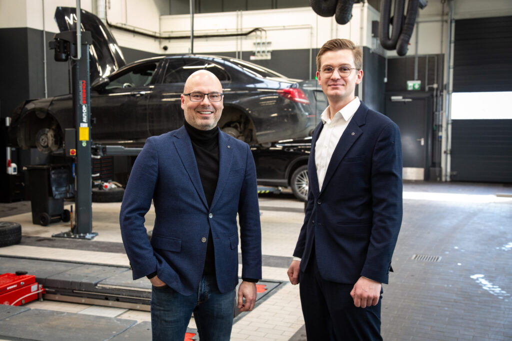 Two men in blue blazers standing in an auto repair shop with a black car on a lift behind them.