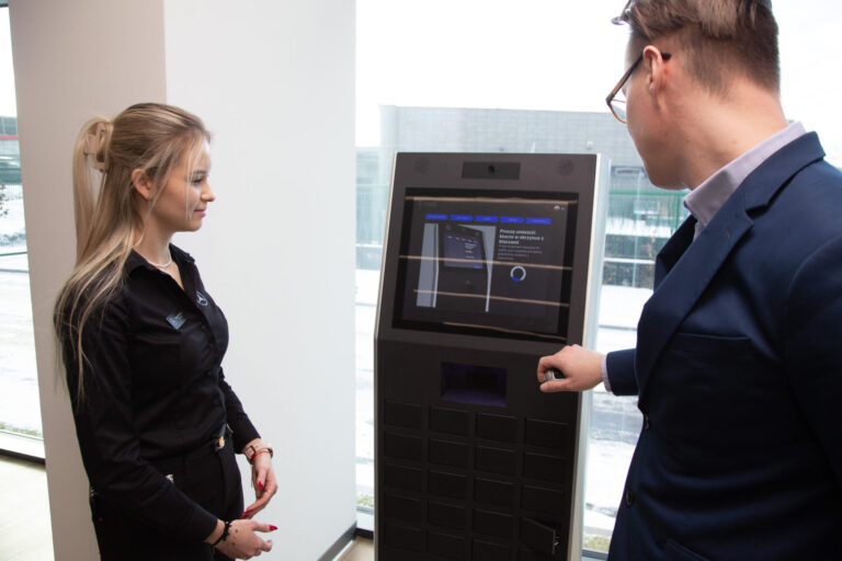 Two people stand beside a tall self-service kiosk in a bright office; a woman on the left watches while a man in a suit operates the screen surface.
