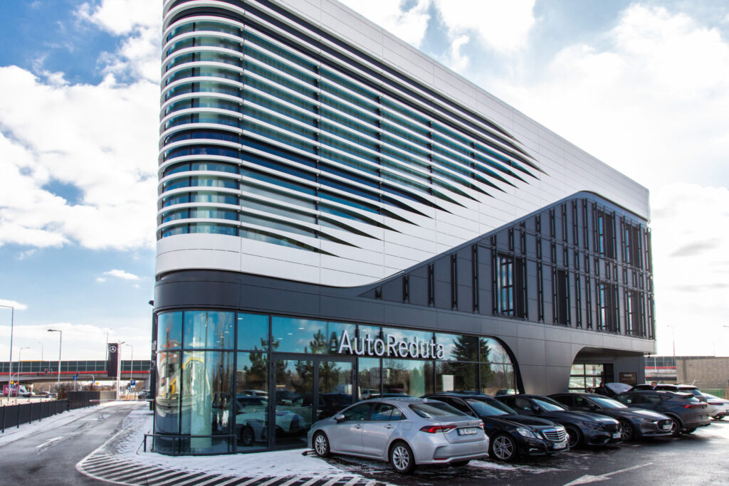 Modern glass and metal building with a curved, striped facade and 'AutoReduta' sign, a car showroom with parked cars in front.