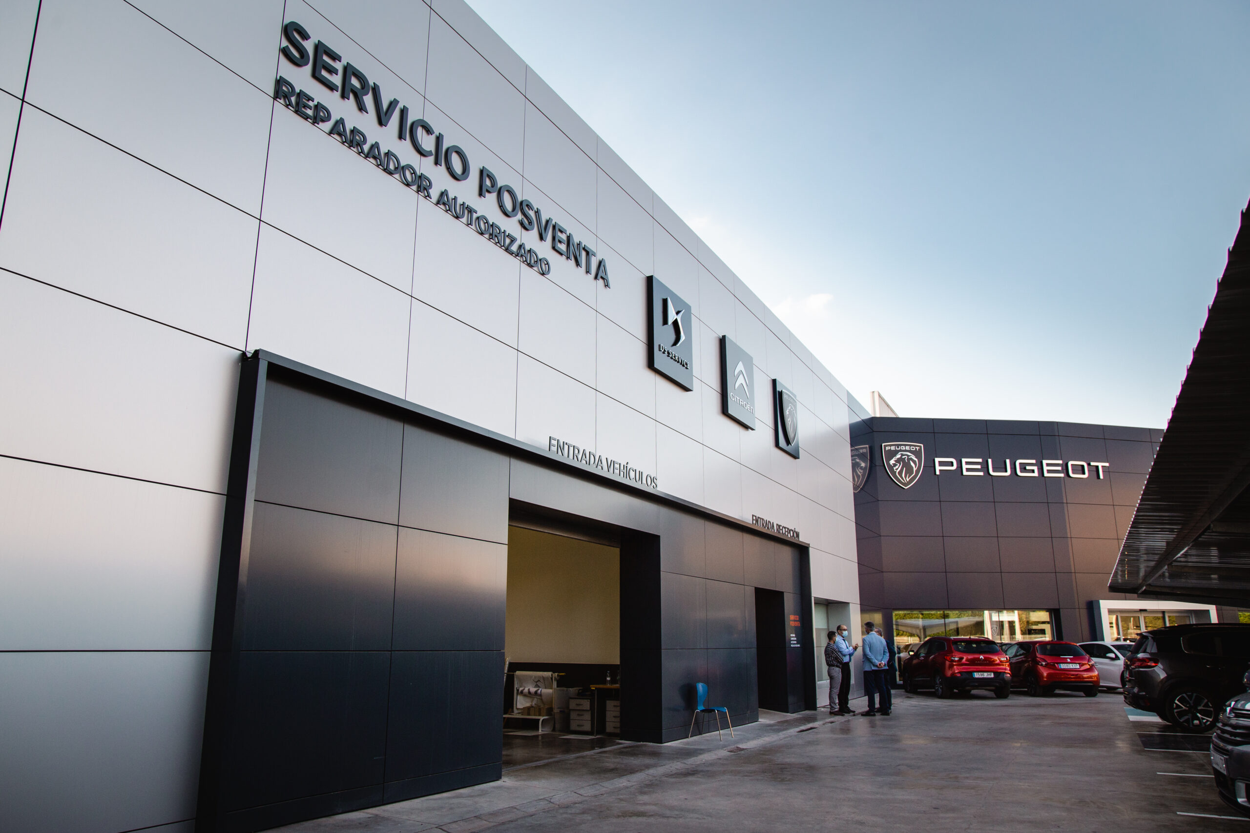 Exterior view of a car service center with a large 'SERVICIO POSVENTA' sign, authorized repair logos, and several cars parked outside