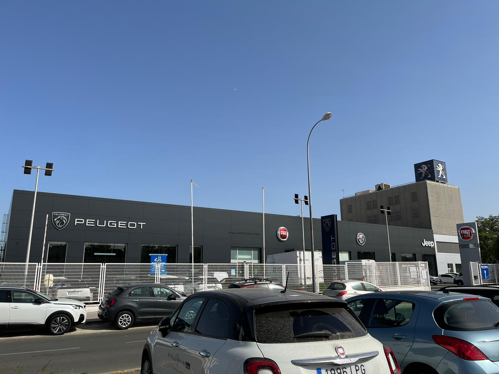 Front of a car dealership with Peugeot signage and logos for Fiat and Jeep under a clear blue sky; rows of parked cars in the lot.