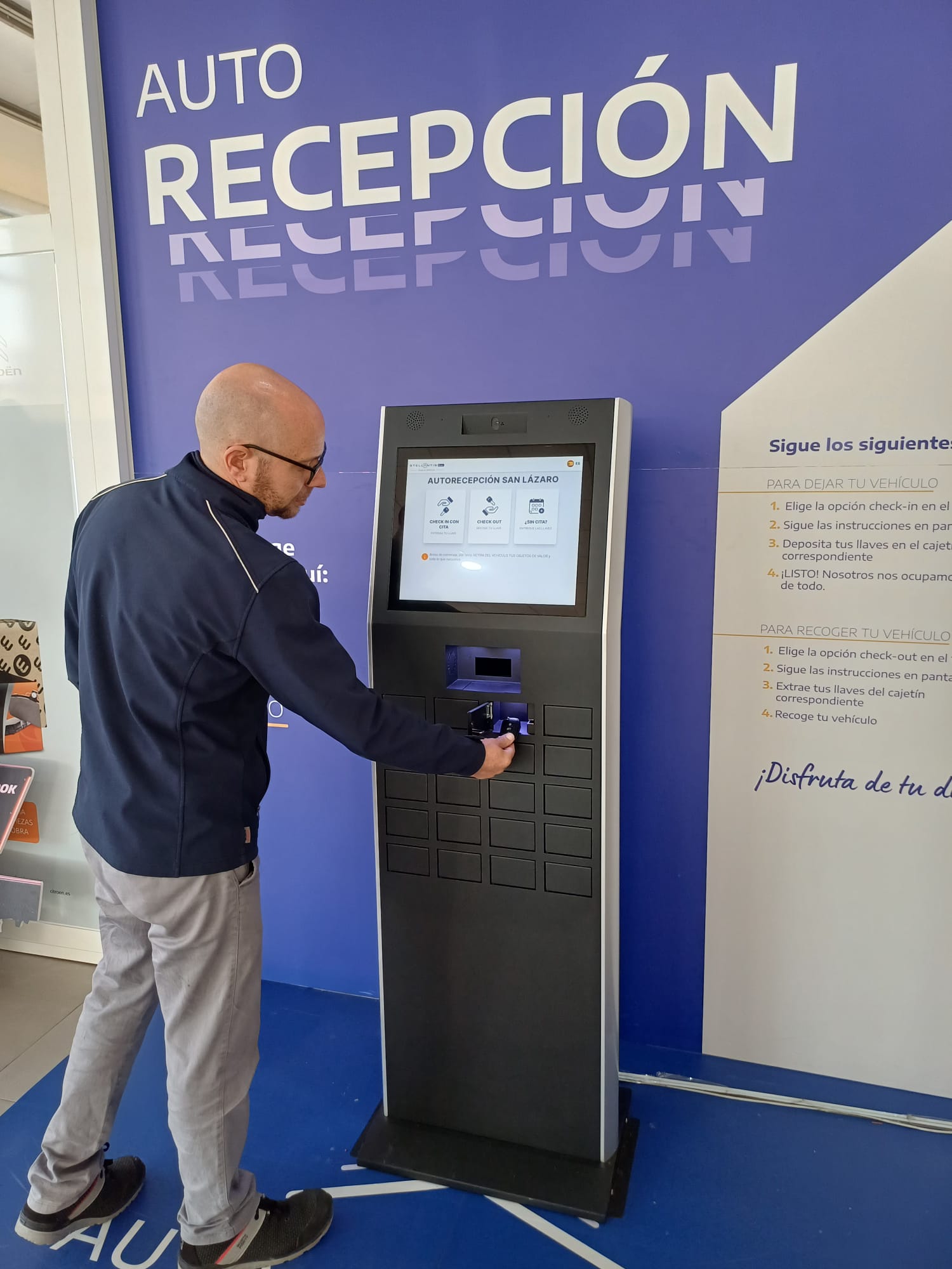 Man using a self-service check-in kiosk in front of a blue wall that reads 'AUTO RECEPCIÓN'.
